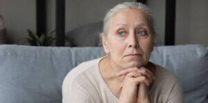 Sad lonely pensive elder lady looking away, sitting on couch at home with chin on hands, thinking over health problems